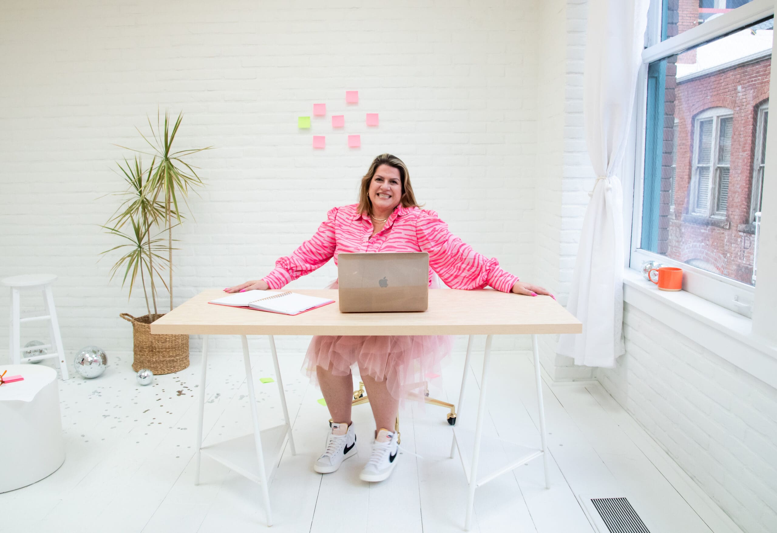 A woman sitting at a desk in a minimal white office, smiling at the camera. A laptop and notebook are on the desk, and she is wearing a pink leopard-print top.